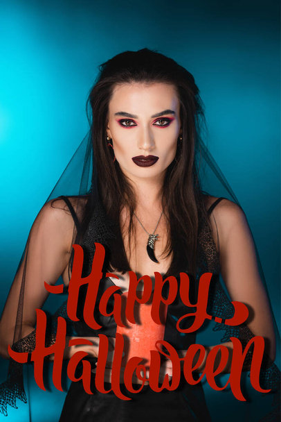 young pale woman with black makeup and veil holding burning candle near happy halloween lettering on blue - Photo, Image