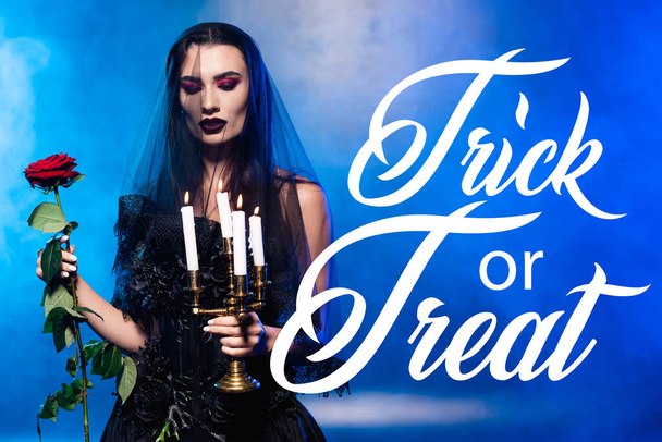 woman in black dress and veil holding red rose and candles near trick or treat lettering on blue with smoke - Foto, imagen
