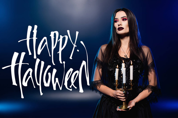 žena v černých šatech a závoj drží hořící svíčky v blízkosti šťastný halloween nápis na modré  - Fotografie, Obrázek