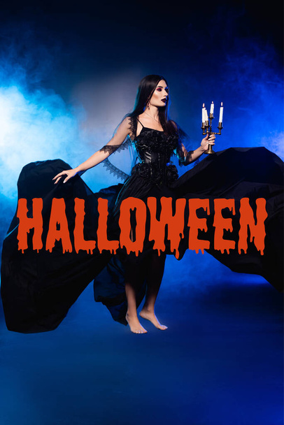 bleke vrouw in zwarte jurk houden kandelaar met brandende kaarsen in de buurt van Halloween belettering op blauw met rook - Foto, afbeelding