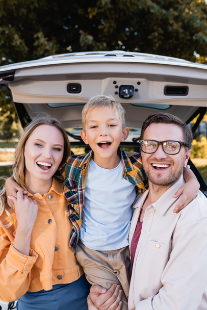 Cheerful boy hugging parents near car during trip outdoors  - Foto, afbeelding