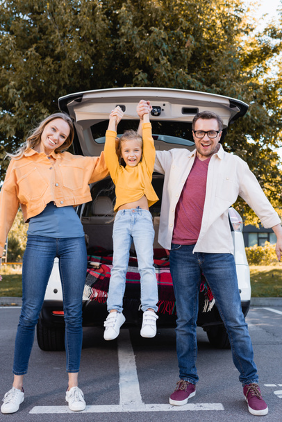 Веселые родители держат дочь возле машины на открытом воздухе  - Фото, изображение