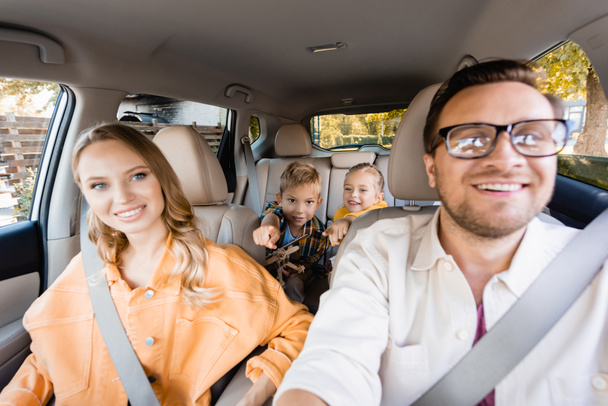 Cheerful kids pointing with fingers near parents on blurred foreground in car  - Photo, Image