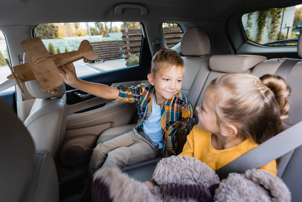 Мальчик с игрушечным самолетом смотрит на сестру с мягкой игрушкой на размытом переднем плане в машине  - Фото, изображение