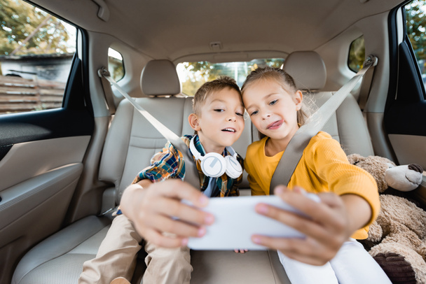 ヘッドフォンやおもちゃを持つ子供たちは、車の中でぼやけた前景にスマートフォンで自撮り  - 写真・画像