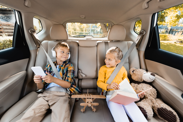 Los niños con teléfono inteligente y libro sonriendo unos a otros cerca de los juguetes en el coche  - Foto, Imagen