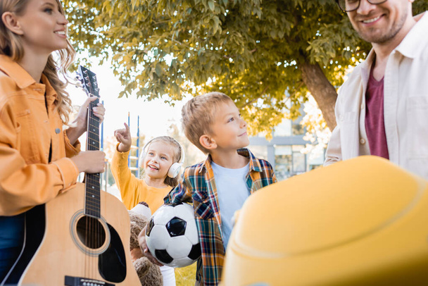 Niños alegres con fútbol y juguete suave de pie cerca de los padres con maleta y guitarra acústica en primer plano borrosa  - Foto, imagen
