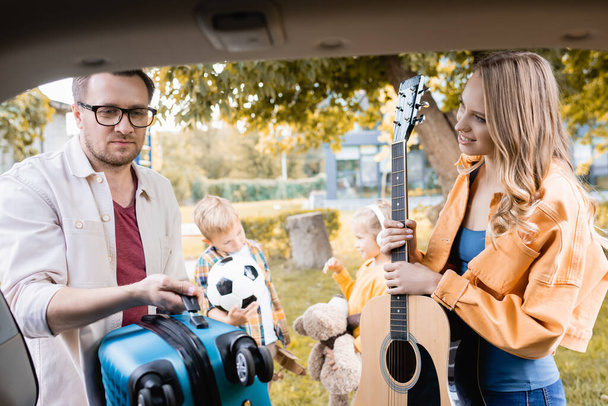 Улыбающаяся женщина с акустической гитарой смотрит на мужа с чемоданом рядом с детьми и багажником автомобиля на размытом переднем плане  - Фото, изображение