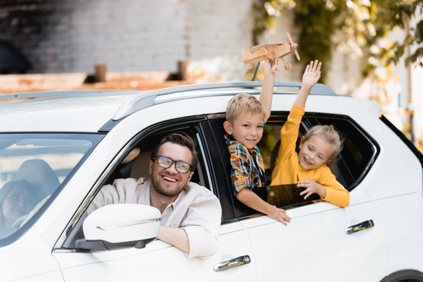 Lächelnder Mann blickt mit Spielzeug im Auto in die Kamera neben Frau und Kindern  - Foto, Bild