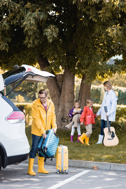 Man in autumn outfit holding suitcase near car and wife with acoustic guitar and kids on blurred background  - Photo, Image