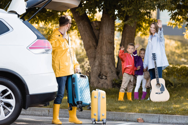 Férfi őszi ruhában integet kezét, miközben gazdaság bőrönd közelében autó és feleség gyerekekkel szabadban  - Fotó, kép