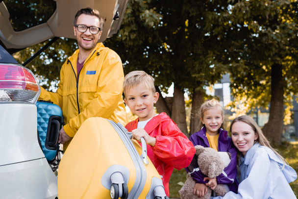 Vrolijke jongen houden koffer in de buurt van familie in de herfst outfit en auto buiten  - Foto, afbeelding