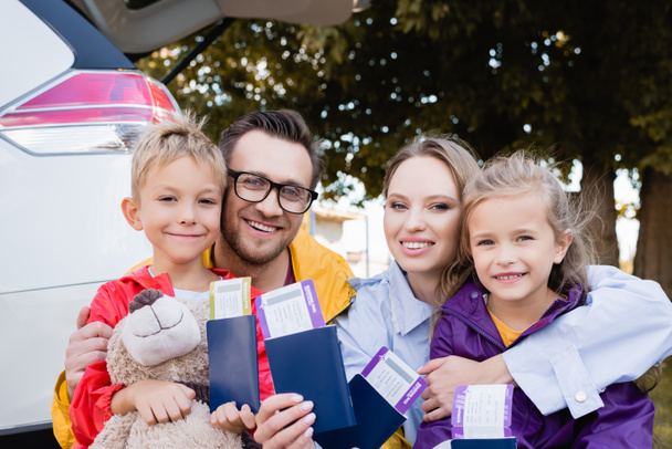 Vrolijke familie in het bezit van paspoorten en vliegtickets in de buurt van auto buiten  - Foto, afbeelding