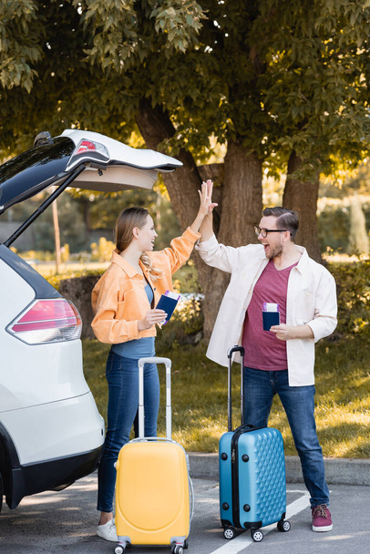 Cheerful couple giving high five while holding passports with air tickets near suitcases and auto  - Photo, Image