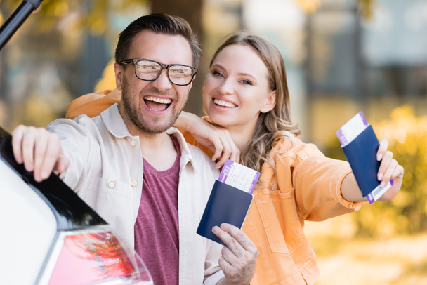 Cheerful woman embracing husband while holding passports with air tickets near car  - Foto, imagen