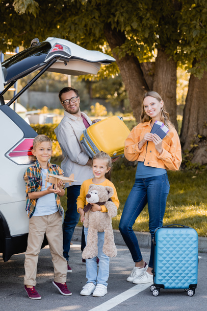 Familia alegre con pasaportes y maletas de pie cerca de auto al aire libre  - Foto, Imagen