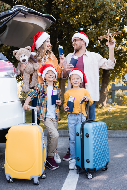 スーツケースの近くに航空券と自動屋外でドキュメントを保持しているサンタの帽子の笑顔の家族  - 写真・画像