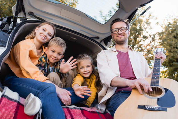 Man holding acoustic guitar near kids waving hands and smiling wife in trunk of car  - Photo, Image