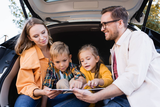 Kinder zeigen auf Karte neben lächelnden Eltern im Kofferraum des Autos  - Foto, Bild