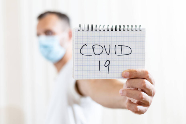 blurred man in a medical mask holds a sheet of paper with the inscription COVID 19 on light background - Photo, Image