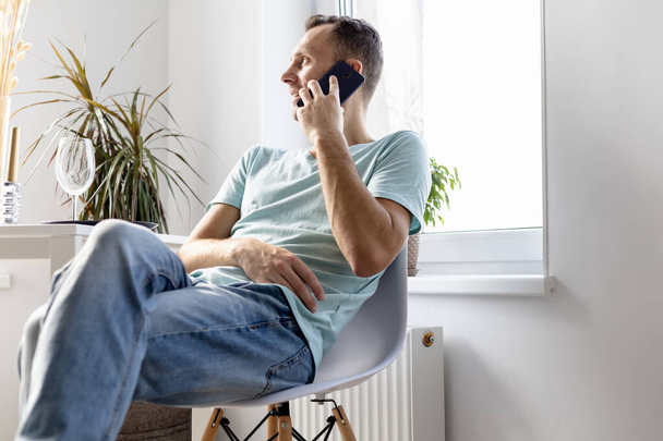 young man in t-shirt and jeans talking on the phone sitting on a chair in a bright room - Foto, imagen