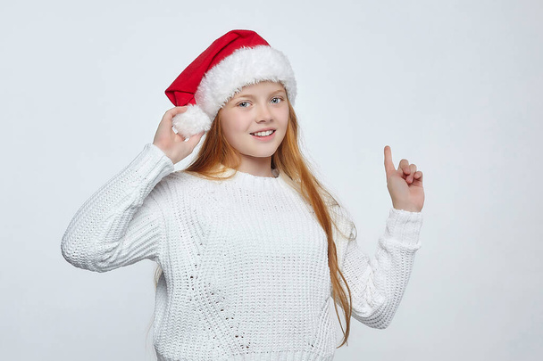 attractive teenage girl with red hair wearing a Santa hat. Indicates with your finger an empty space for text. photo session in the studio on a white background - Foto, immagini