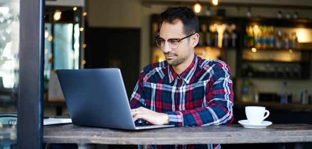 Задумчивый мужчина-фрилансер в повседневной одежде и очках сидит за столом в кафе и работает над удаленным проектом на ноутбуке - Фото, изображение