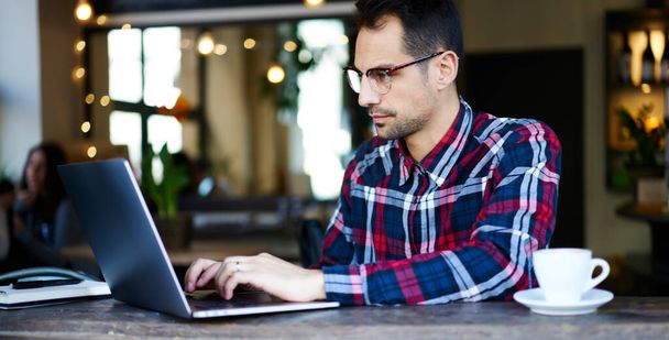 Pensive bearded male freelancer in casual wear and glasses sitting at wooden table in cozy cafe and working on project - Photo, Image