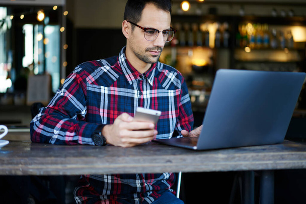 Adult brunette man looking at laptop screen and text messaging on mobile phone while having break in cozy workspace in modern cafe - Photo, Image