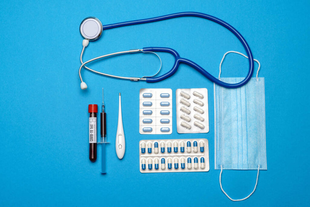 prevention and treatment of coronavirus healthcare concept - syringe, mask, tablets, stethoscope and Covid-19 test tube on blue background - Foto, afbeelding