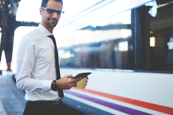 Smiling adult office worker in formal clothing and eyeglasses standing on railway platform and waiting for train while using smartphone and having coffee to go - 写真・画像