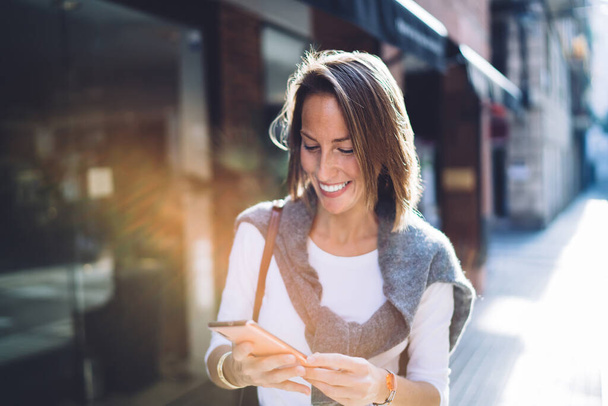 Smiling caucasian woman reading news from social networks holding mobile phone walking on street, positive female influencer strolling with smartphone sending mail and messages during sunny day - Foto, Imagen