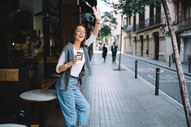 Щаслива молода жінка в повсякденному одязі посміхається і тримає чашку гарячої кави, стоячи на вулиці і махаючи рукою
 - Фото, зображення