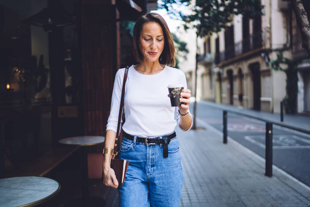 Good looking middle aged female in casual wear walking on pavement with notebook in hand and enjoying hot coffee to go - Foto, Imagem