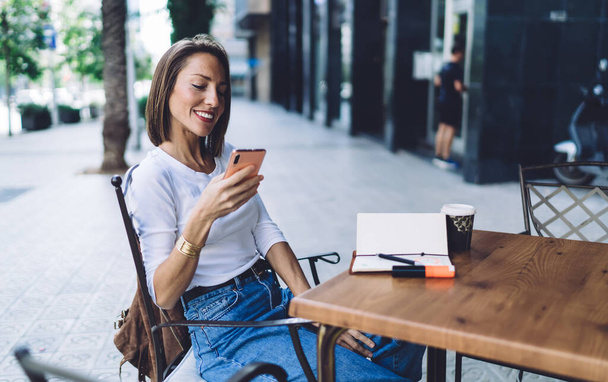 Happy female entrepreneur in white shirt and blue jeans reading text message and browsing internet on phone while enjoying coffee break in summer cafe - Φωτογραφία, εικόνα