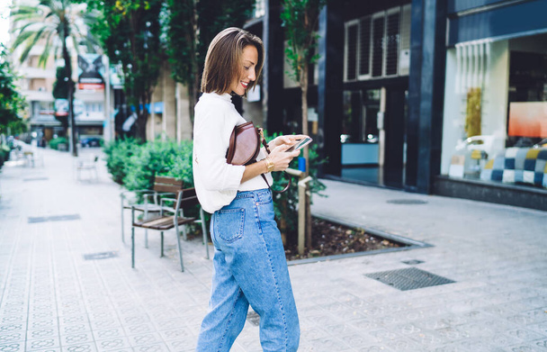 Side view of happy adult female in stylish casual outfit walking along street and looking at phone while browsing internet - Фото, зображення