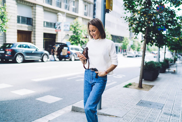 Happy lady in casual outfit using mobile on street while walking hand in pocket on modern city street on sunny day - 写真・画像