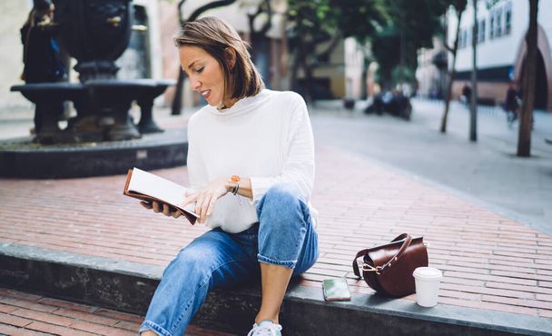 Ajattelevainen 40-vuotias nainen trendikkäässä asussa lukemassa tietoja muistikirjasta istuessaan kiviportailla puistossa kahvitauon aikana. - Valokuva, kuva