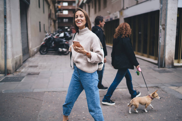 Positive female pedestrian in jeans and sweater with backpack and smartphone walking in downtown against blurred urban background and looking at camera - Fotografie, Obrázek