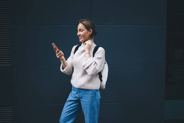 Smiling adult female in casual clothes with backpack standing near blue wall and browsing mobile phone while looking at screen - Zdjęcie, obraz