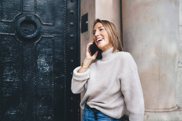 Cheerful adult female in casual clothes with toothy smile and standing near entrance of old building while talking on mobile phone - Foto, Imagen