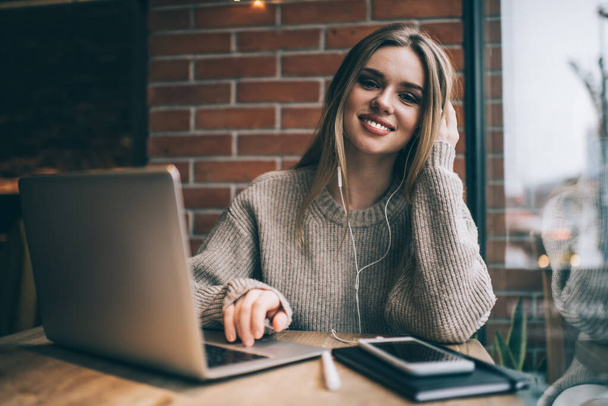 Young happy female with long blond hair in knitted sweater smiling and working with laptop while sitting at table near window and looking at camera - Photo, Image