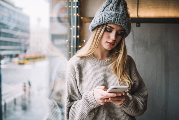 Millennial blogger using modern cellular gadget for social networking and web chatting, female generation Z dressed in knitted pullover and winter hat connecting to 4g wireless on mobile technology - Fotografie, Obrázek