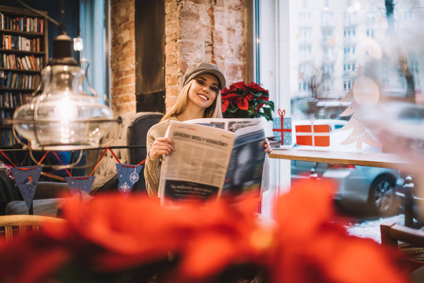 Portrait of cheerful hipster girl in trendy cap holding journal with daily news and smiling at camera during rest time in loft cafeteria with X-mas decoration, happy Caucasian woman with newspaper - Photo, Image