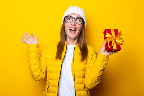 Surprised joyful young woman in yellow jacket holding a gift on yellow background - Valokuva, kuva