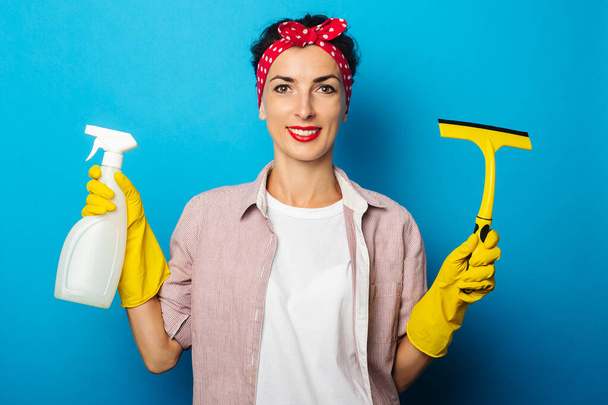 Uśmiechnięta młoda kobieta w rękawiczkach ze sprayem i skrobakiem do czyszczenia okien na niebieskim tle - Zdjęcie, obraz