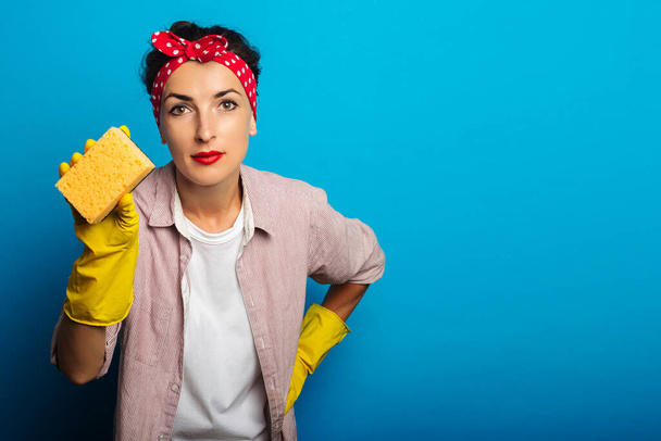Young woman in a bandage for hair, in yellow gloves holding a sponge on a blue background - Фото, изображение