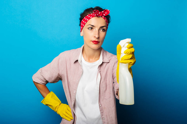 Young woman in cleaning gloves holding spray looks at it on blue background - 写真・画像