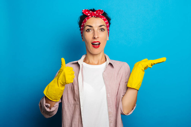 Surprised young woman in gloves holding an invisible object showing thumb up gesture on blue background - Photo, Image