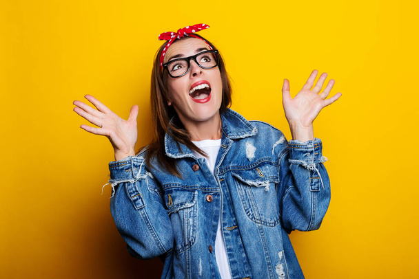 Young woman in a hair band, in a denim jacket joyfully looks up on a yellow background - 写真・画像